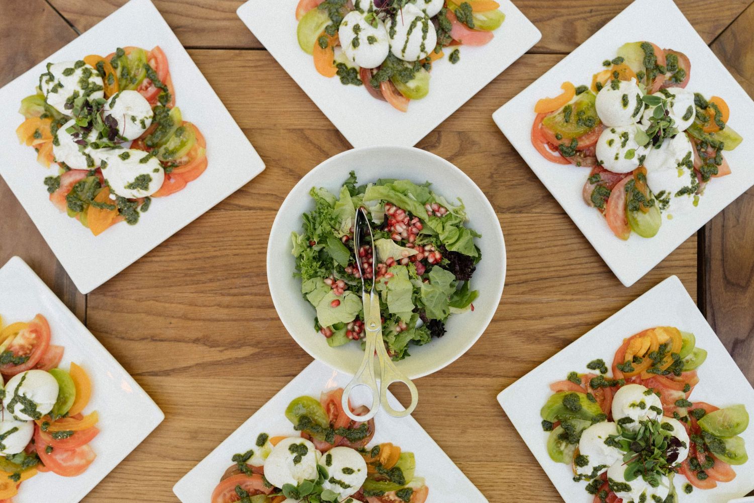 Overhead view of caprese salad plates with burrata, tomatoes, and greens on wooden table.