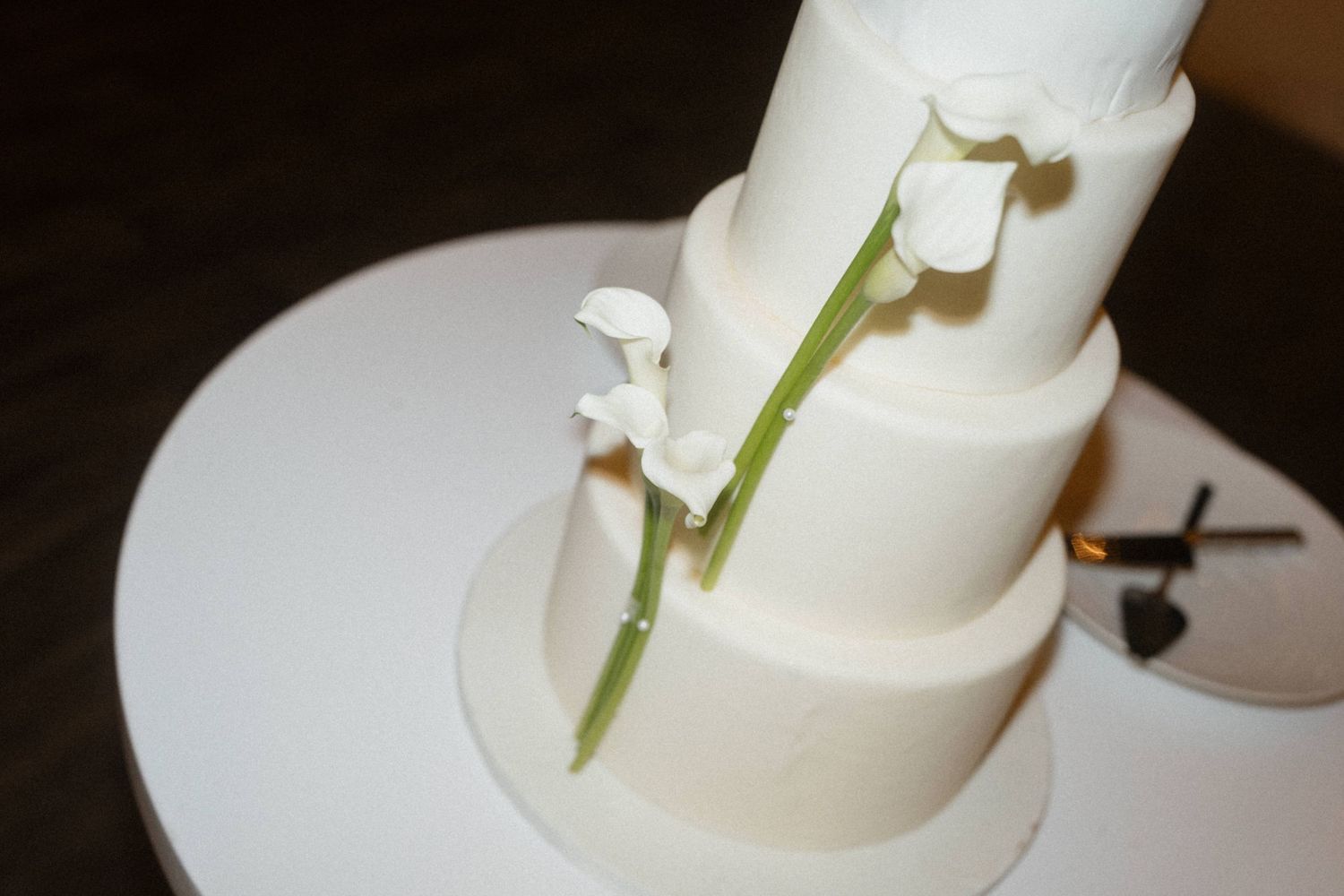 Elegant white tiered wedding cake decorated with fresh calla lily stems on a white cake stand.