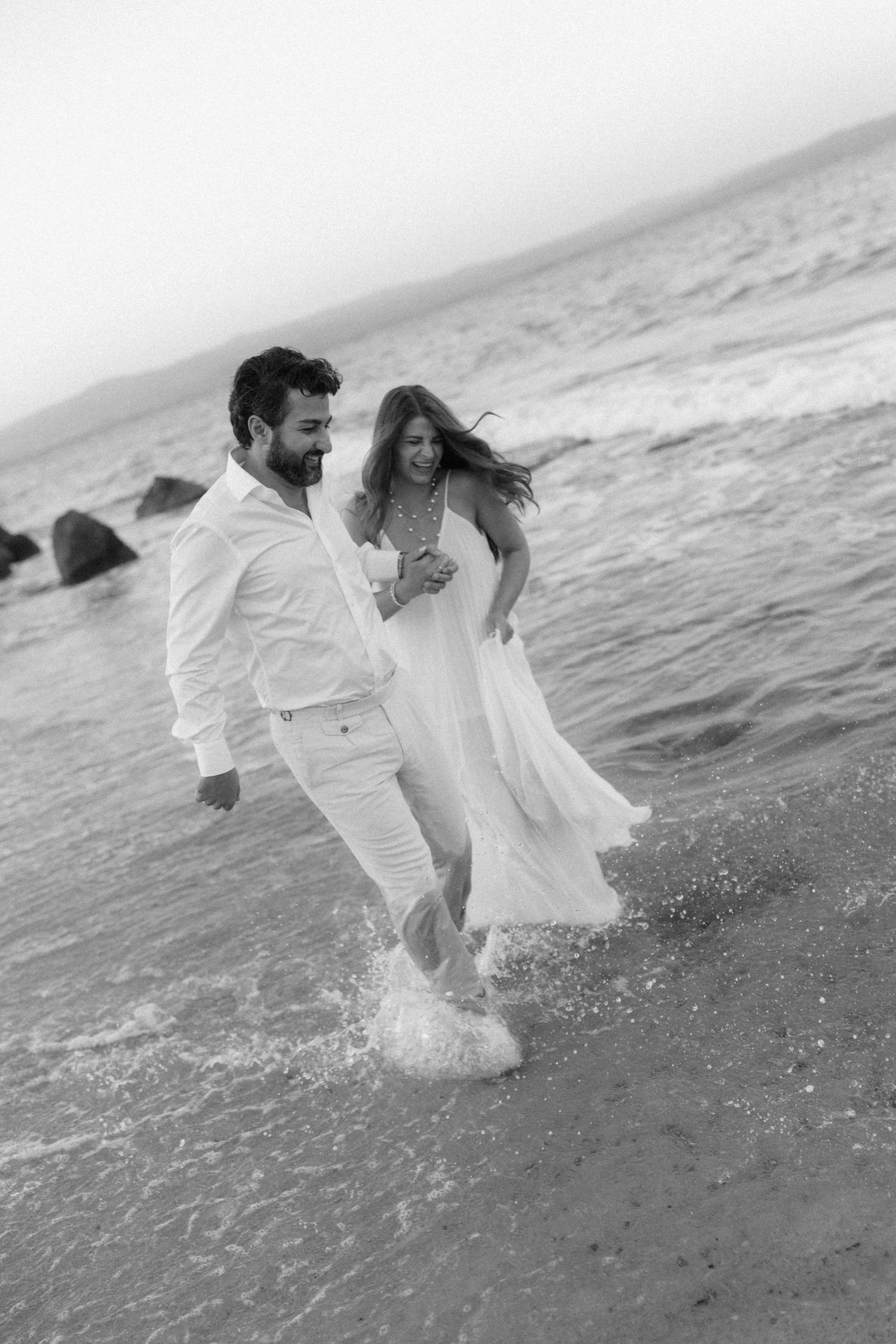 Black and white photo of joyful couple in white running through ocean waves on a rocky beach.
