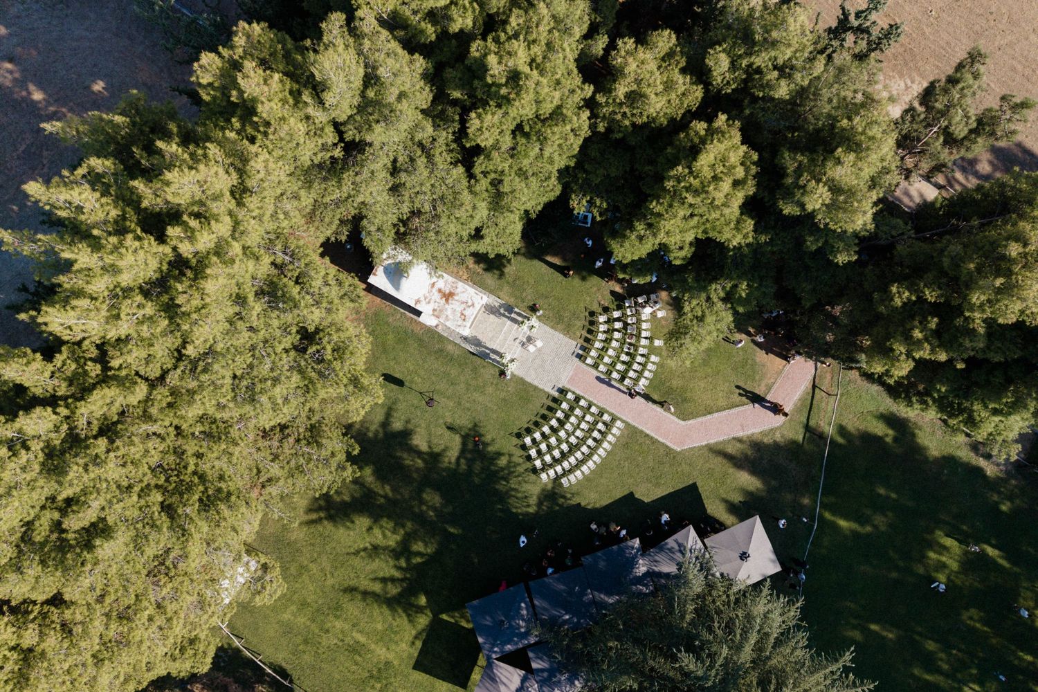 Aerial view of an outdoor wedding ceremony setup with white chairs on a lawn surrounded by tall green trees.