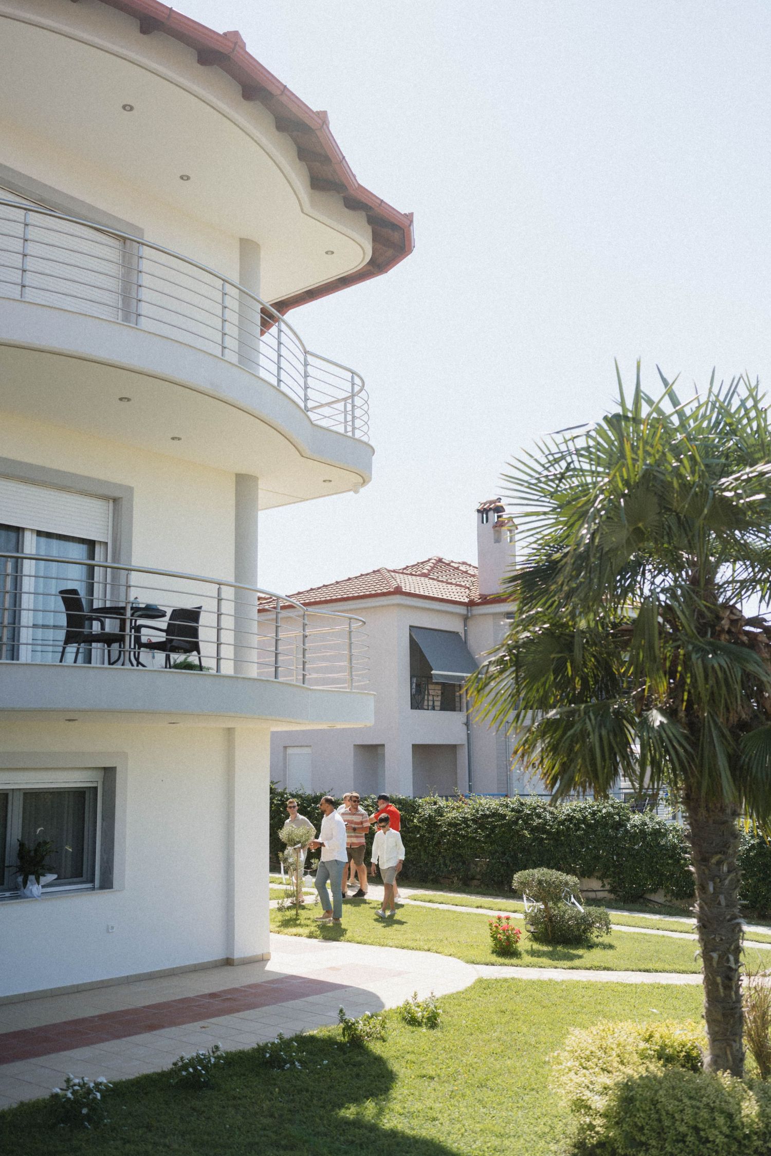 Modern white multi-story villa with balconies, palm trees, and people socializing in a lush green garden.