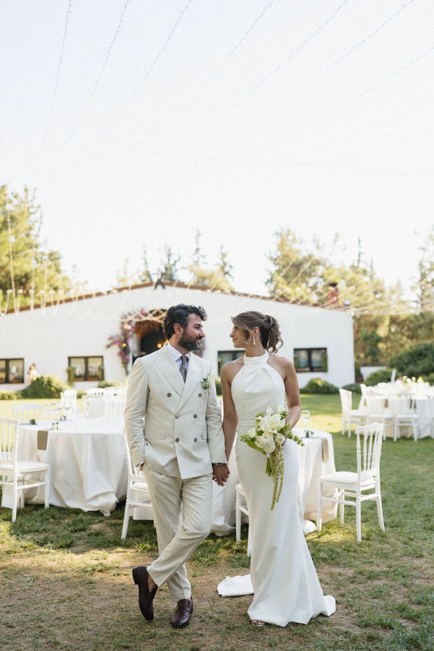 Bride in white halter gown and groom in beige suit walk hand-in-hand at an outdoor garden wedding reception.