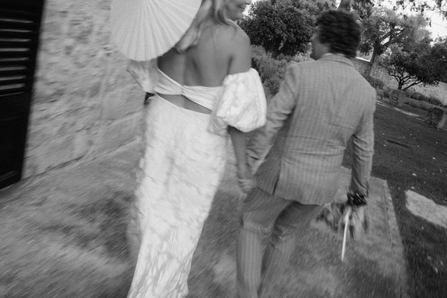 Black and white photo of a bride in a lace gown and groom walking along a rustic stone building at their wedding.