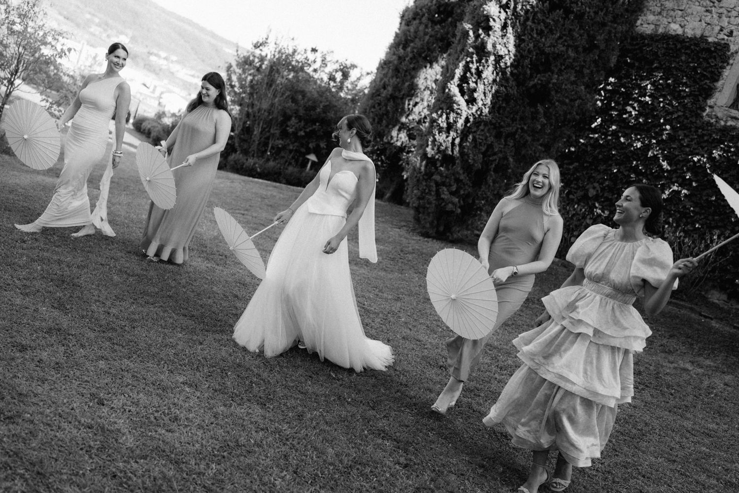 Bride and bridesmaids laughing and walking outdoors with parasols in elegant gowns, captured in black and white.