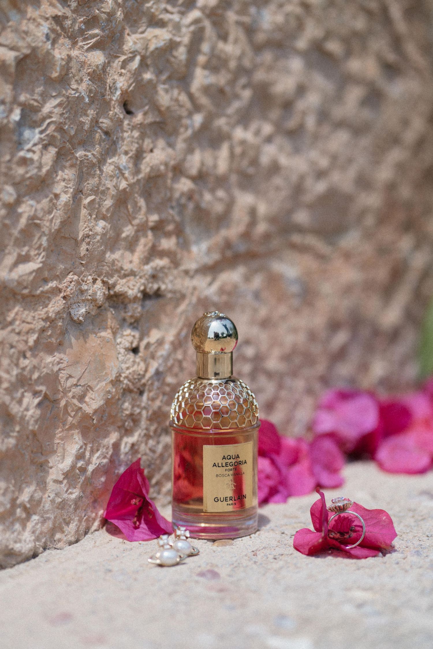 Guerlain Aqua Allegoria perfume bottle with pink flower petals and pearl earrings on stone surface.