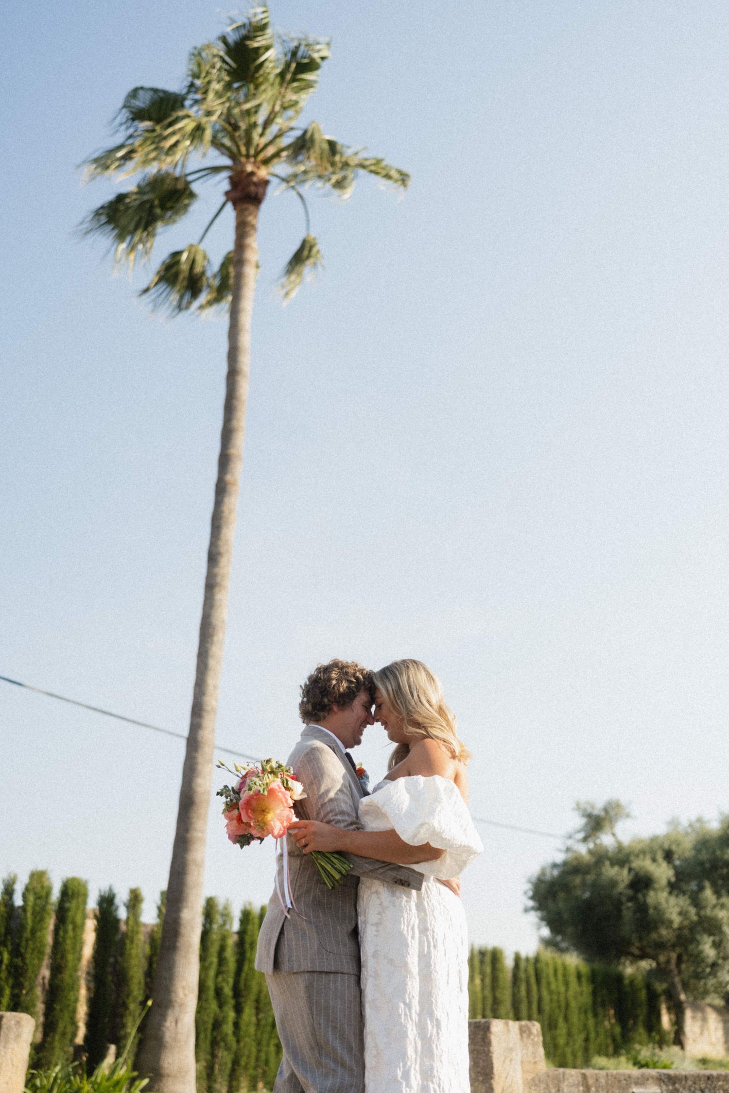 Bride and groom embrace beneath a tall palm tree, holding a coral bouquet against a clear blue sky.