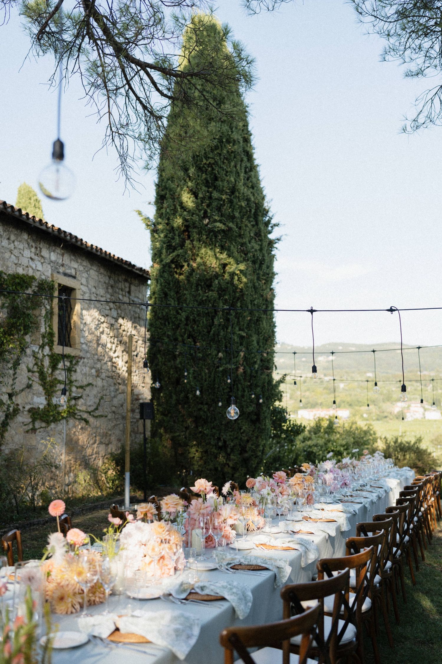 Elegant outdoor wedding reception table with floral centerpieces, string lights, and a cypress tree backdrop at a rustic stone villa.