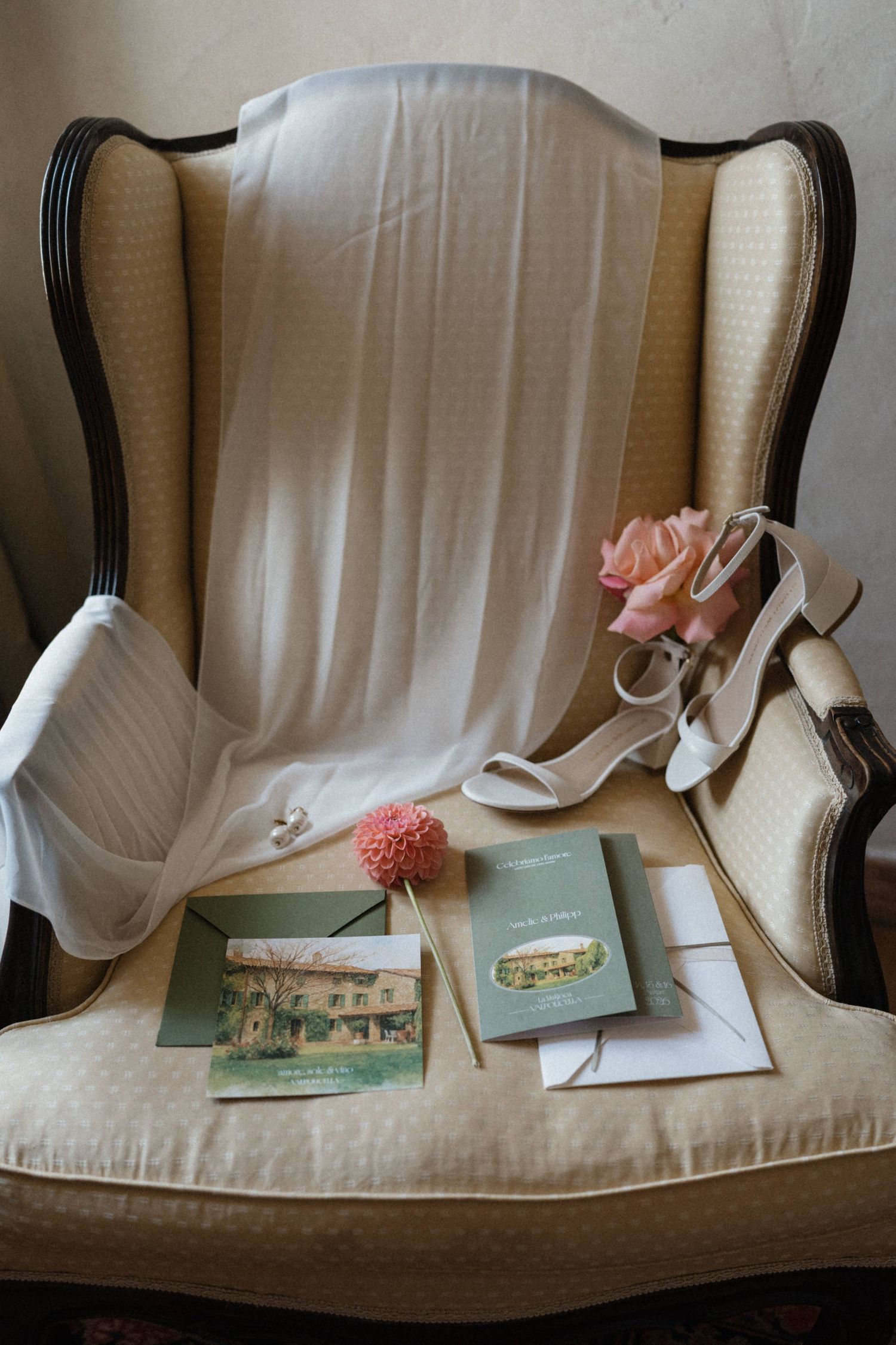 Elegant wedding flat lay with white heels, pink roses, green stationery cards, and bridal veil on a vintage cream wingback chair.
