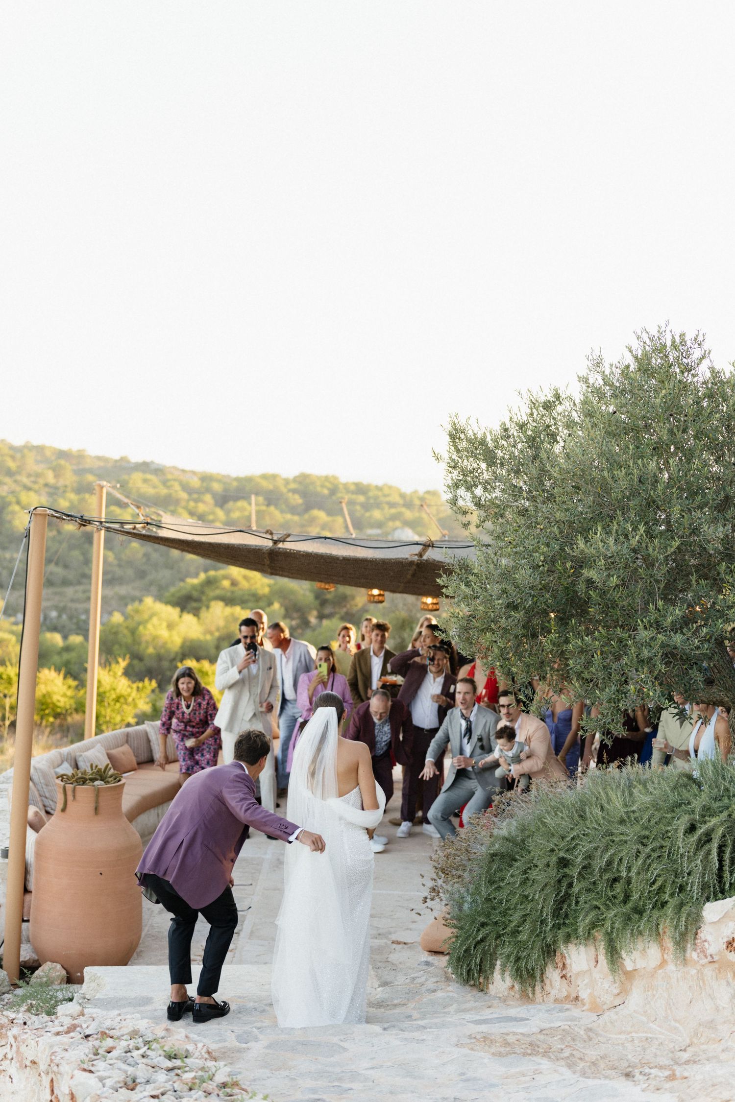 Bride in a long white dress and veil walks down a stone path toward cheering wedding guests beneath an olive tree.