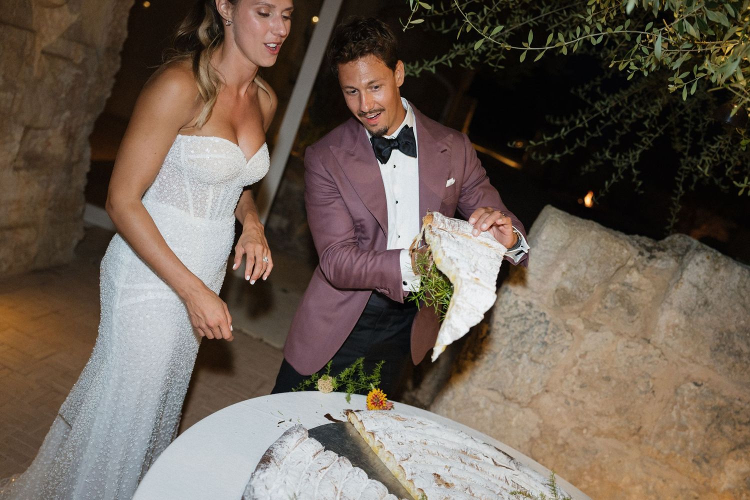 Smiling bride in a strapless lace gown and groom in a burgundy blazer and bow tie cut a rustic wedding cake outdoors at night.