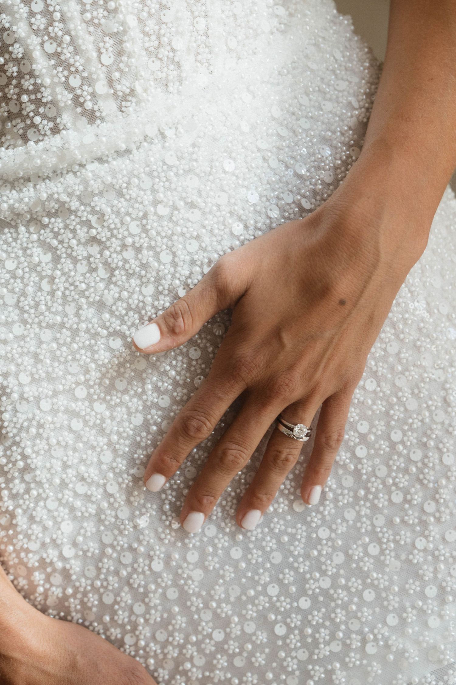 Bride's hand with a diamond engagement ring and white manicure resting on a beaded pearl wedding dress.