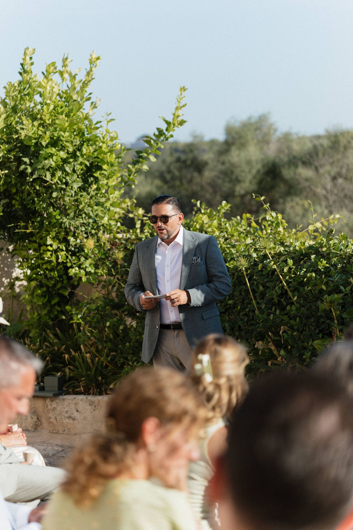 Man in a gray blazer and sunglasses gives a speech to seated wedding guests in a green garden setting.