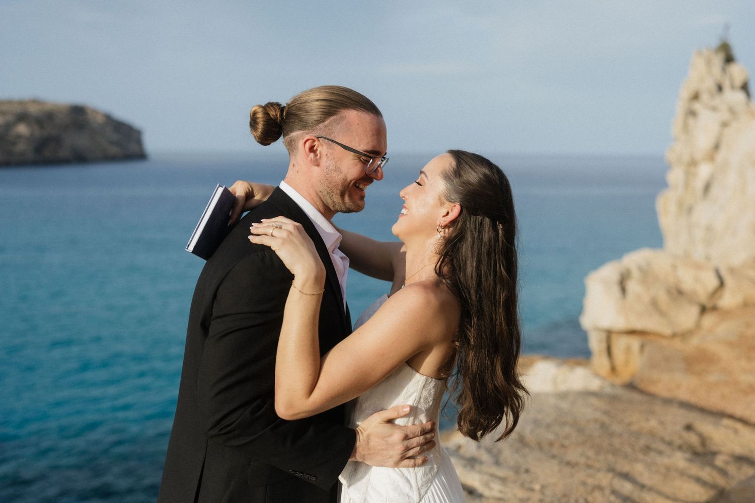 Smiling bride and groom with a man bun embrace on a coastal cliff, with turquoise sea and rock formations behind them.
