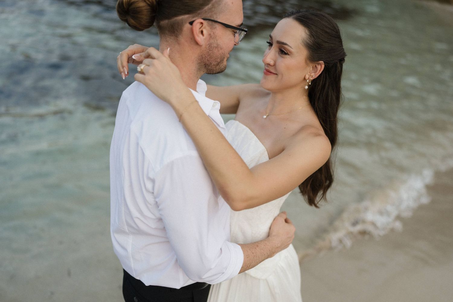 Couple in white embracing on a sandy beach as gentle waves roll in, the woman gazing off to the side in a romantic moment.