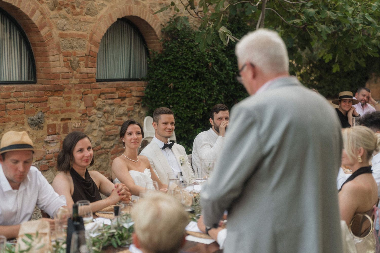 Wedding guests smile as an elderly man gives a speech at an outdoor reception near a rustic brick venue.