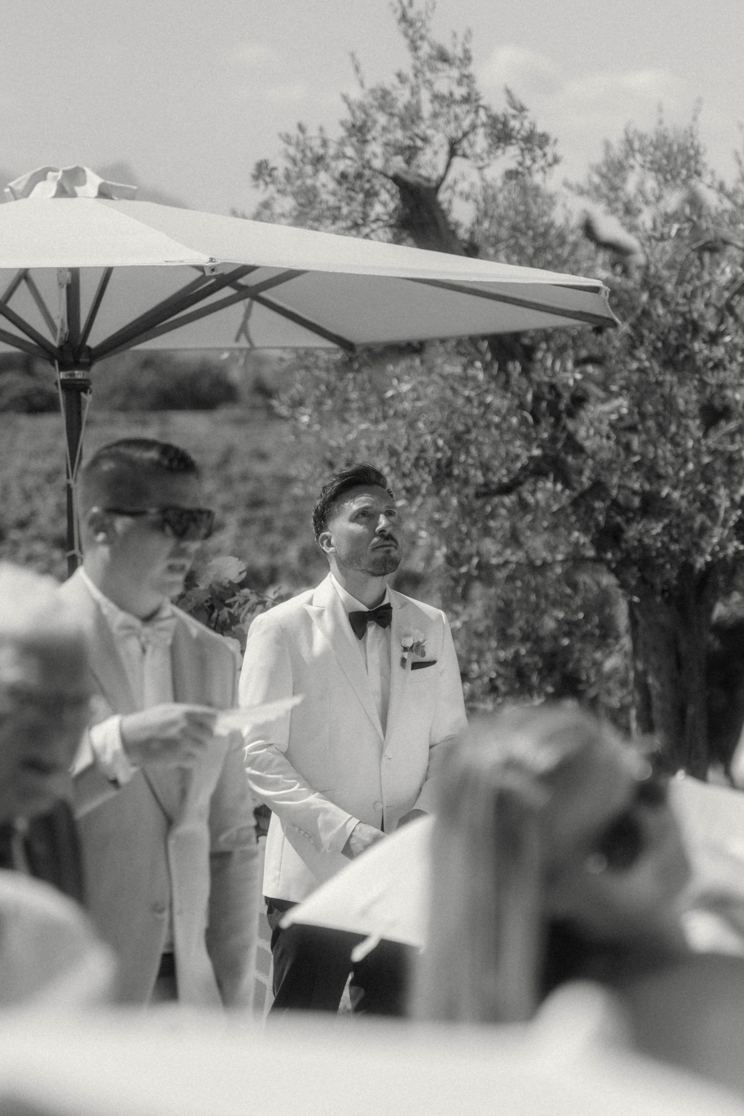 Groom in white tuxedo jacket and bow tie waiting at outdoor wedding ceremony.