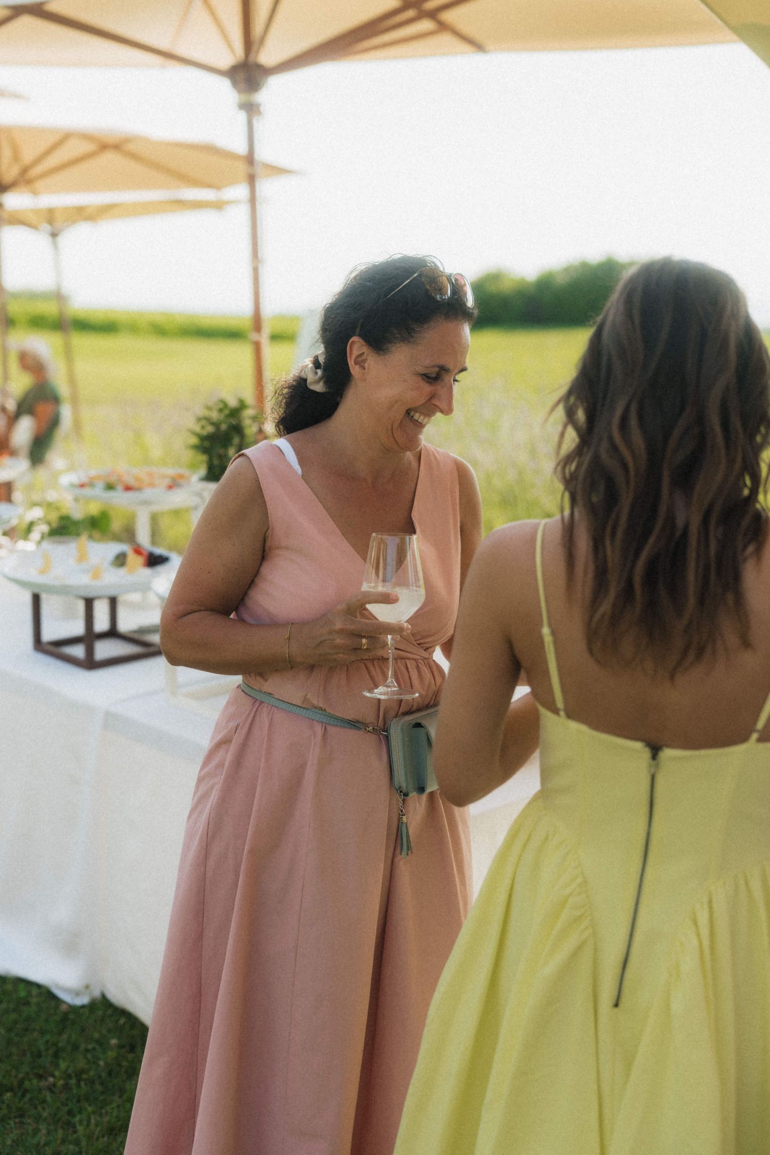 Wedding guest in pink dress holding wine glass chatting with woman in yellow dress.