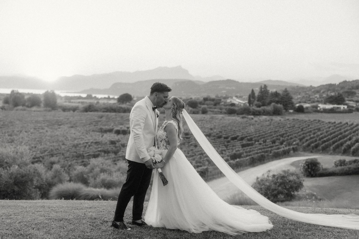 Black and white photo of bride and groom kissing in a vineyard with mountains and a lake in the background.