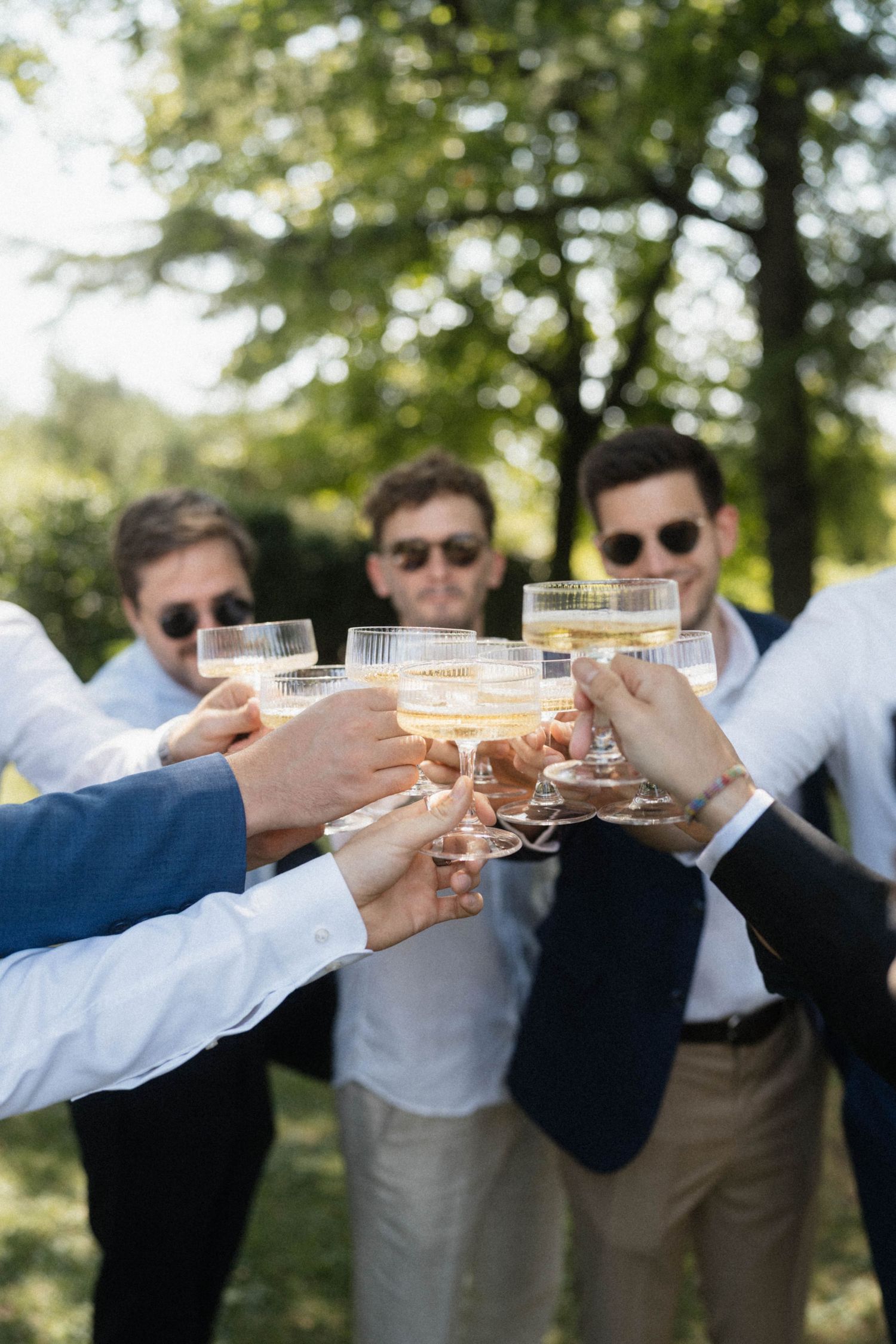 Group of groomsmen in sunglasses toasting with champagne glasses outdoors at sunny wedding celebration.