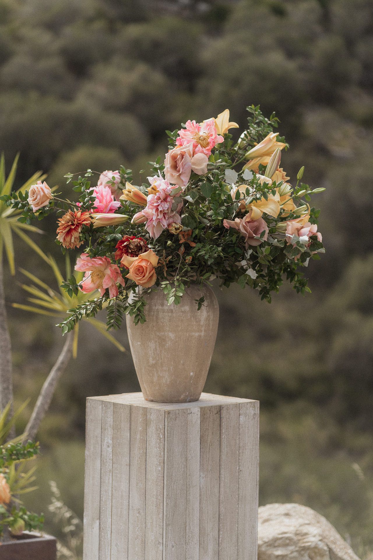 A rustic stone vase filled with pink roses, lilies, and greenery sits atop a white pedestal outdoors.