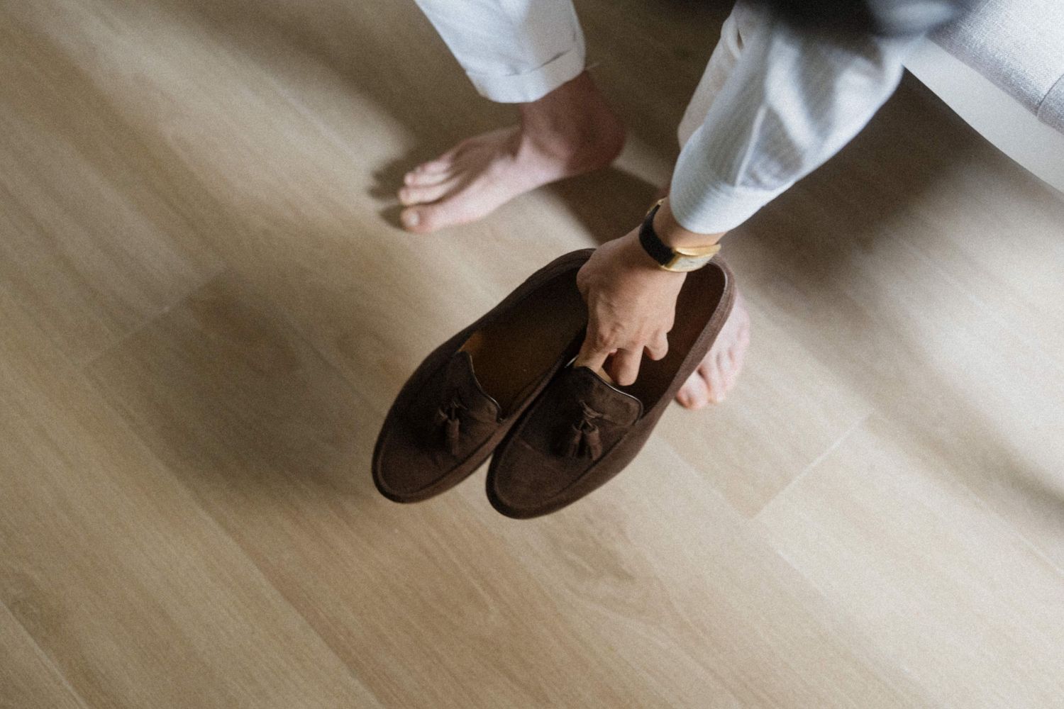 Groom holding brown suede tassel loafers while standing on light wood flooring in white trousers.