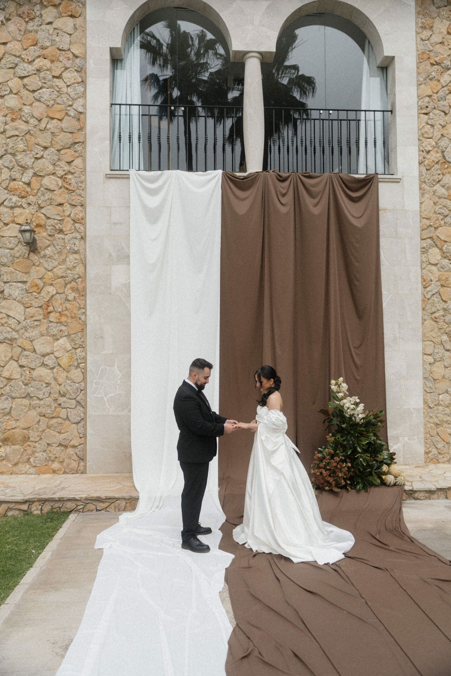 Wedding couple exchanging vows in front of white and brown fabric backdrop at stone building venue.
