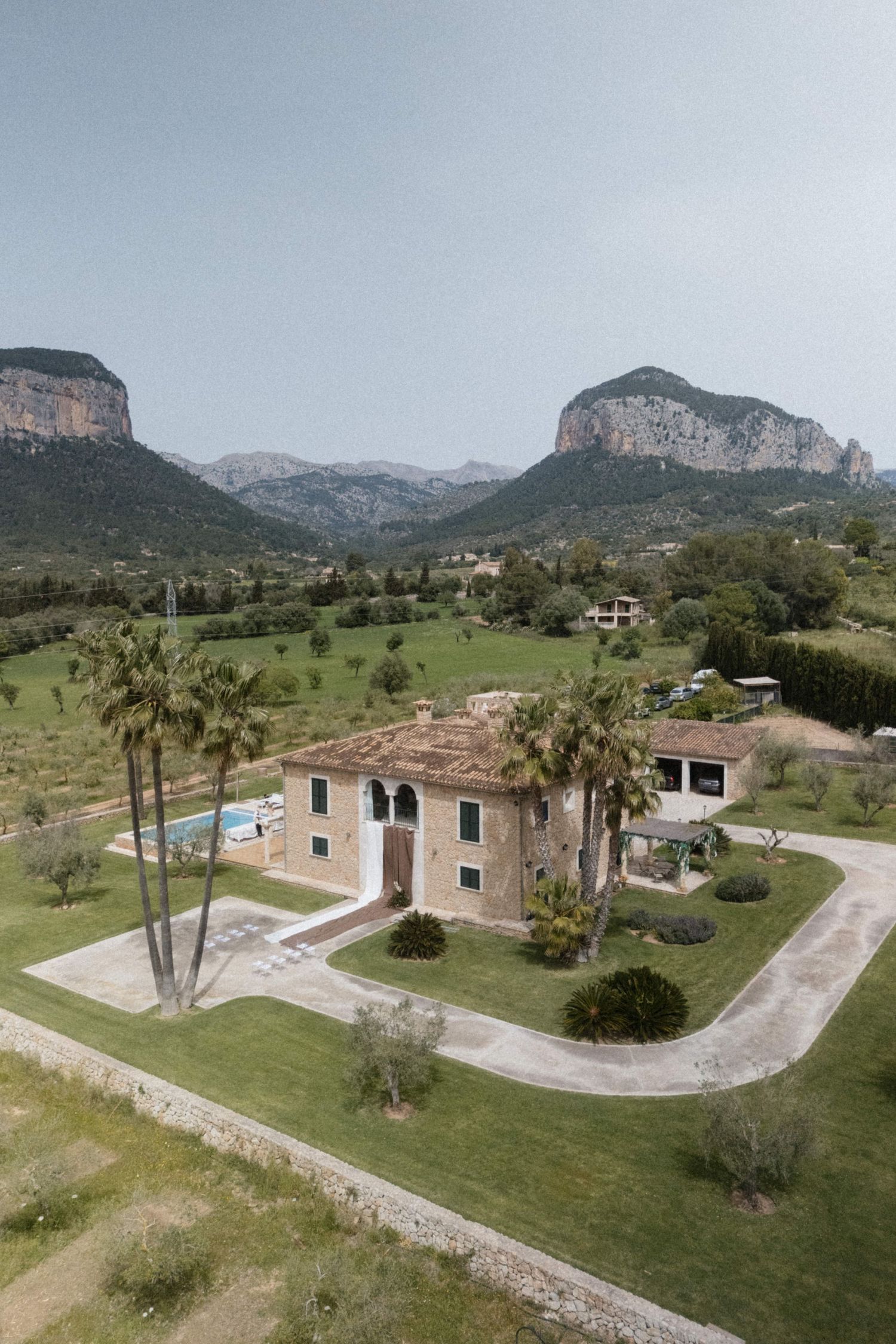 Aerial view of a rustic stone villa with palm trees, pool, and lush green grounds backed by dramatic mountains.