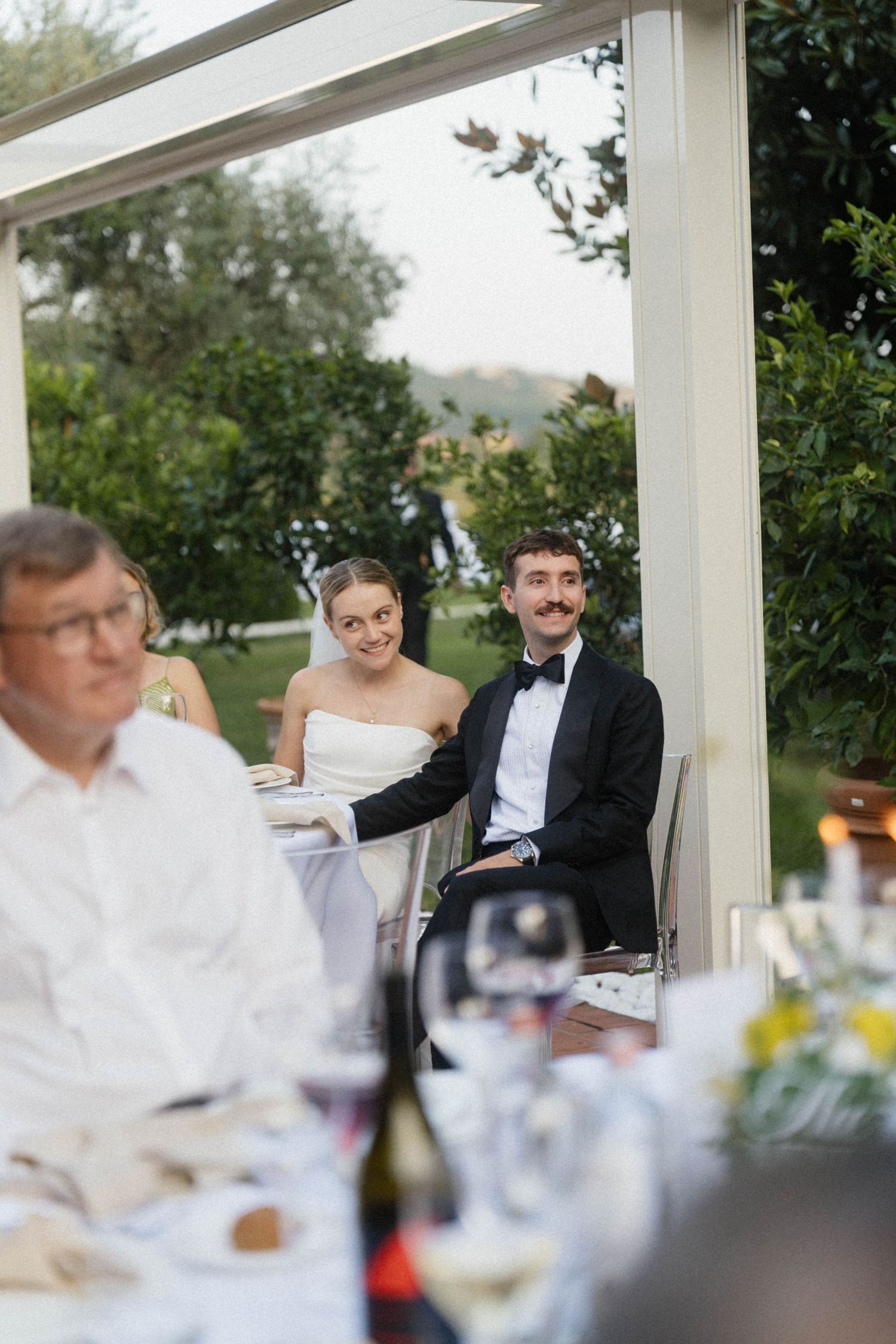 Bride and groom sitting together at outdoor wedding reception table, smiling at guests under white canopy.