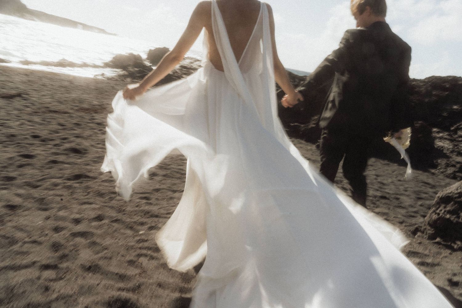 Bride in flowing white backless gown runs with groom on dark sandy beach.