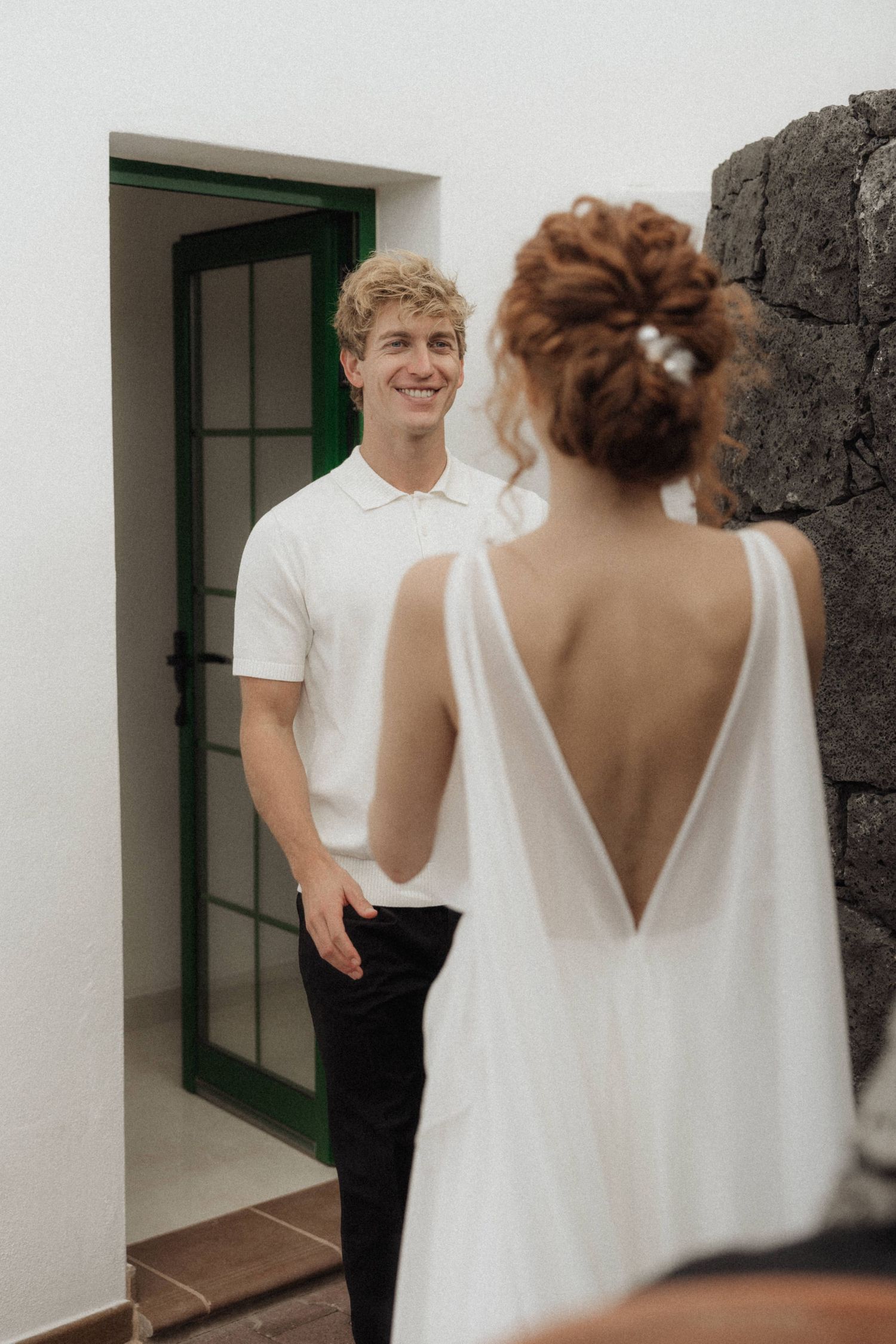Groom in white polo smiles at bride in backless wedding dress during first look moment.