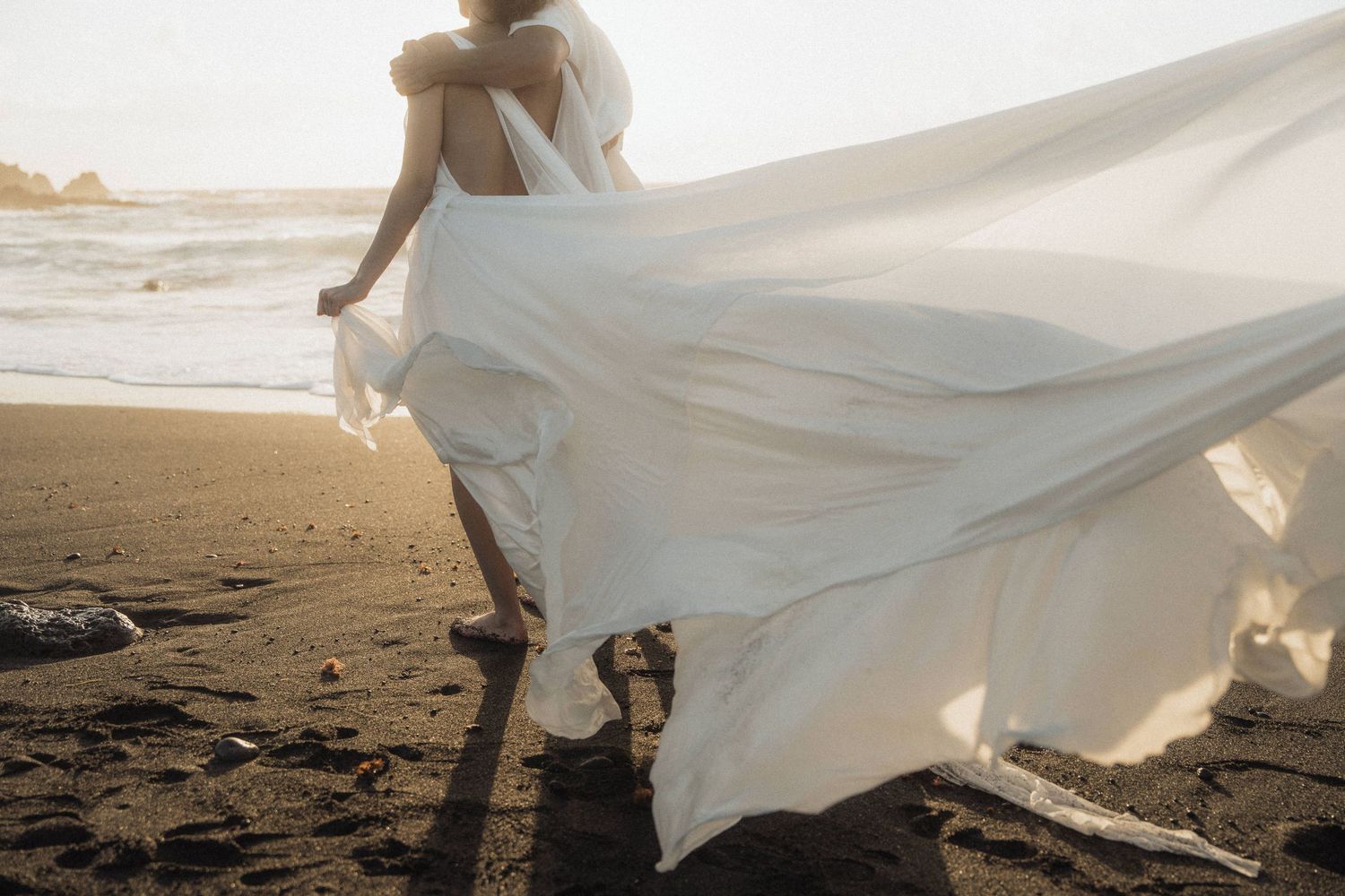 Bride walks along a dark sandy beach as her flowing white wedding dress trails dramatically in the wind.