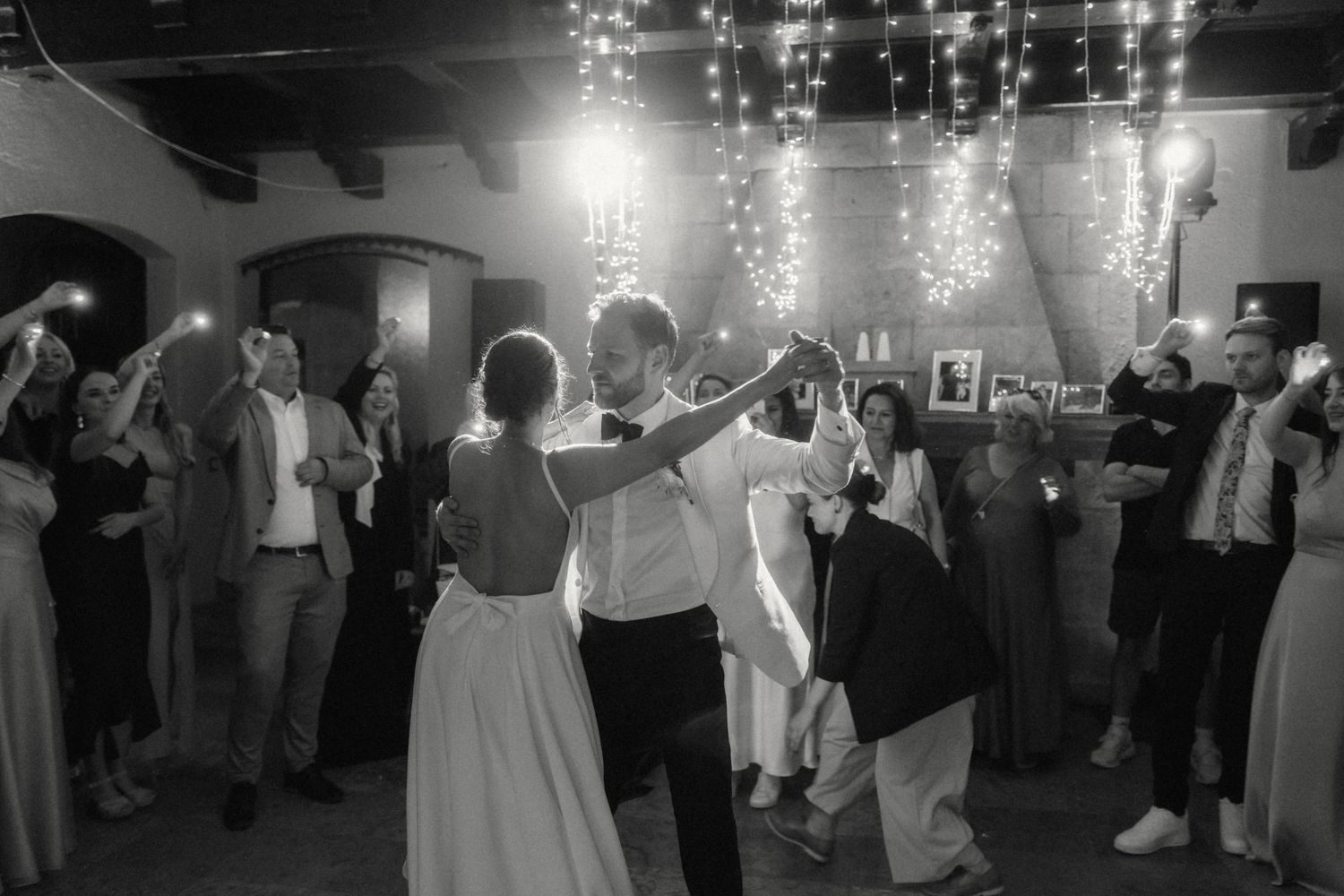 Black and white photo of bride and groom sharing first dance surrounded by celebrating guests.
