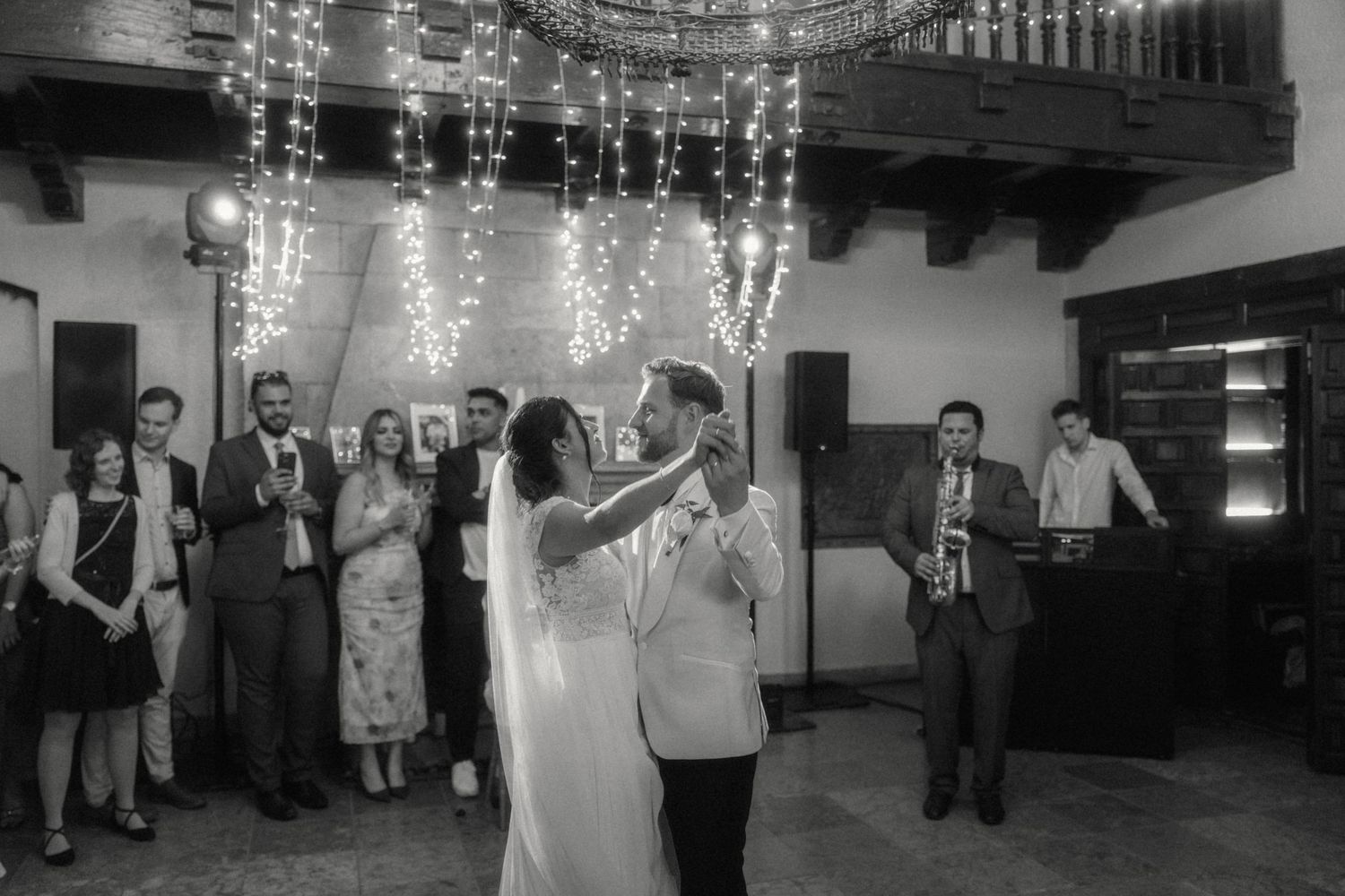 Black and white photo of bride and groom sharing first dance under fairy lights with live saxophone band and guests.