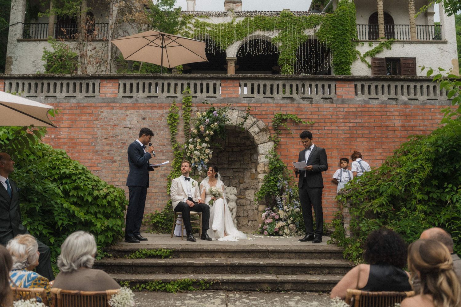 Outdoor wedding ceremony on stone steps with couple seated before officiant, surrounded by lush greenery.