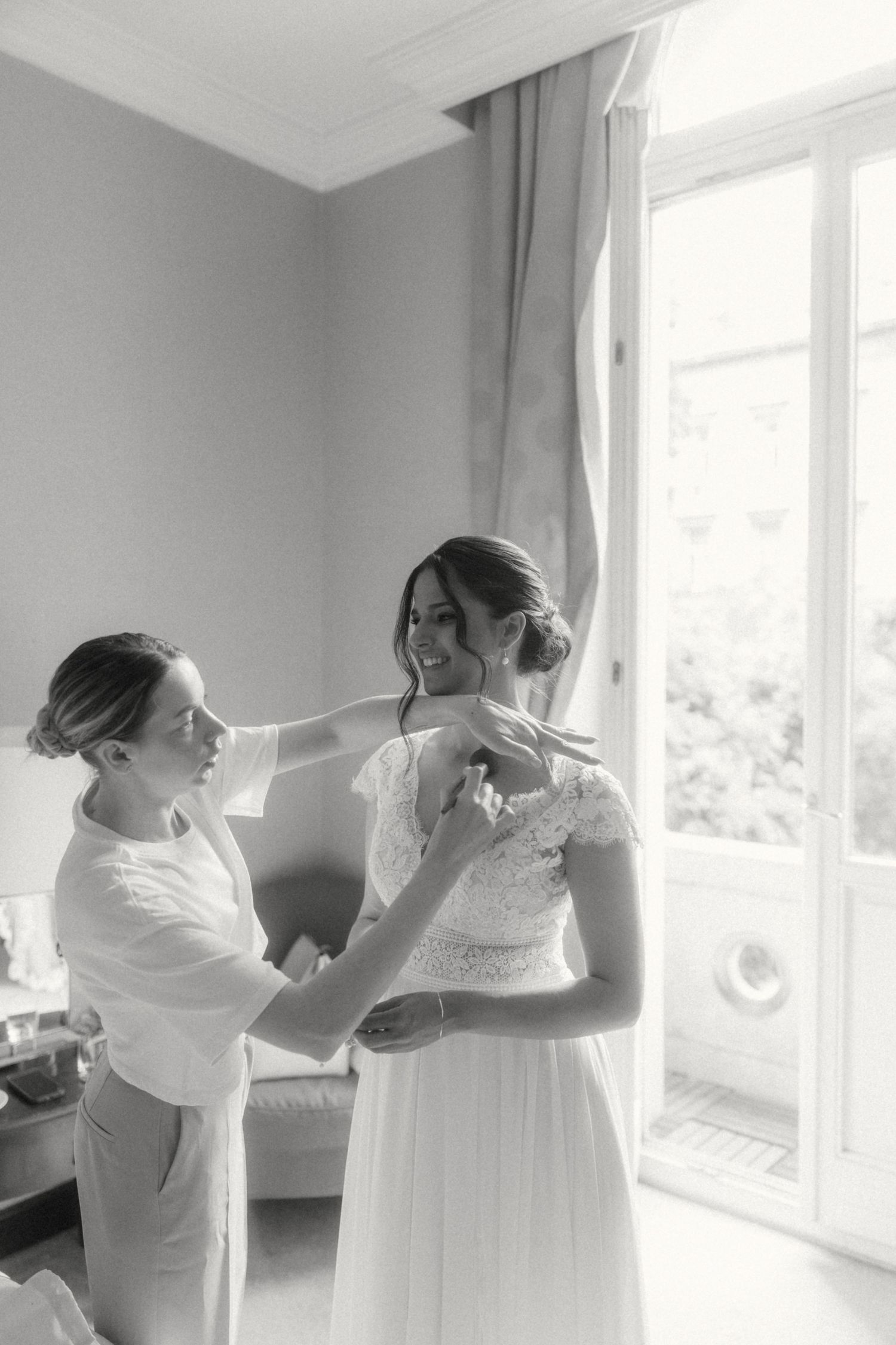 Black and white photo of bridesmaid helping smiling bride button her lace wedding dress by a bright window.
