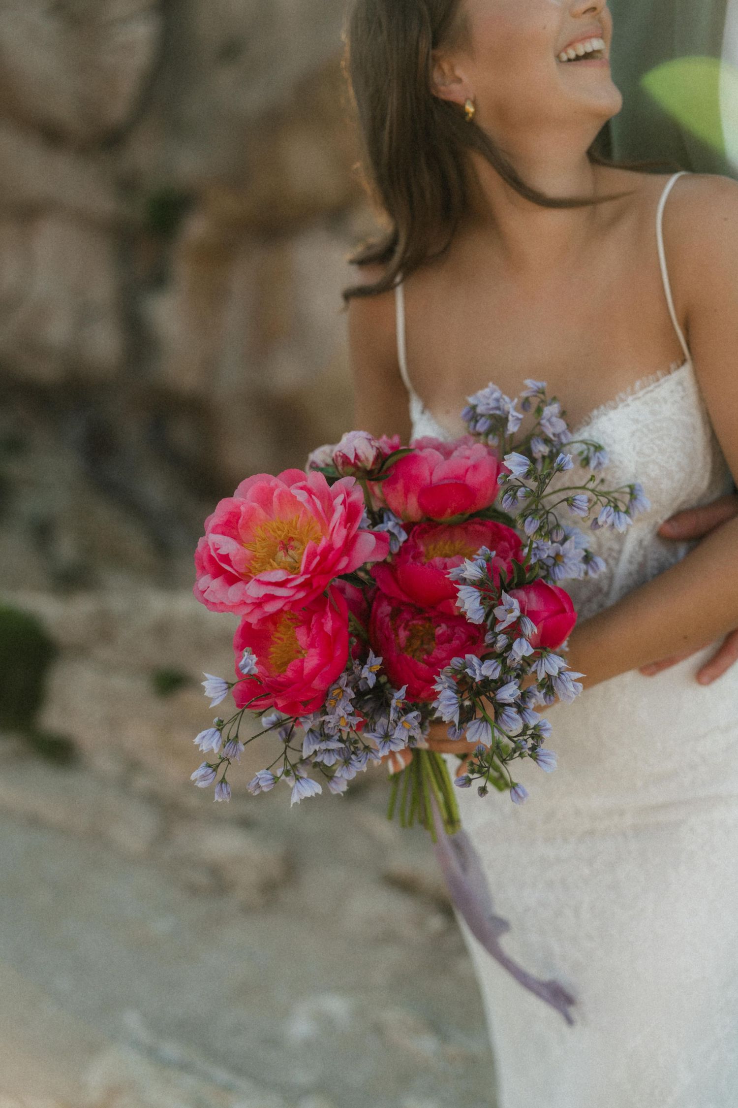 Smiling bride in white lace dress holding vibrant pink peony and lavender wedding bouquet.