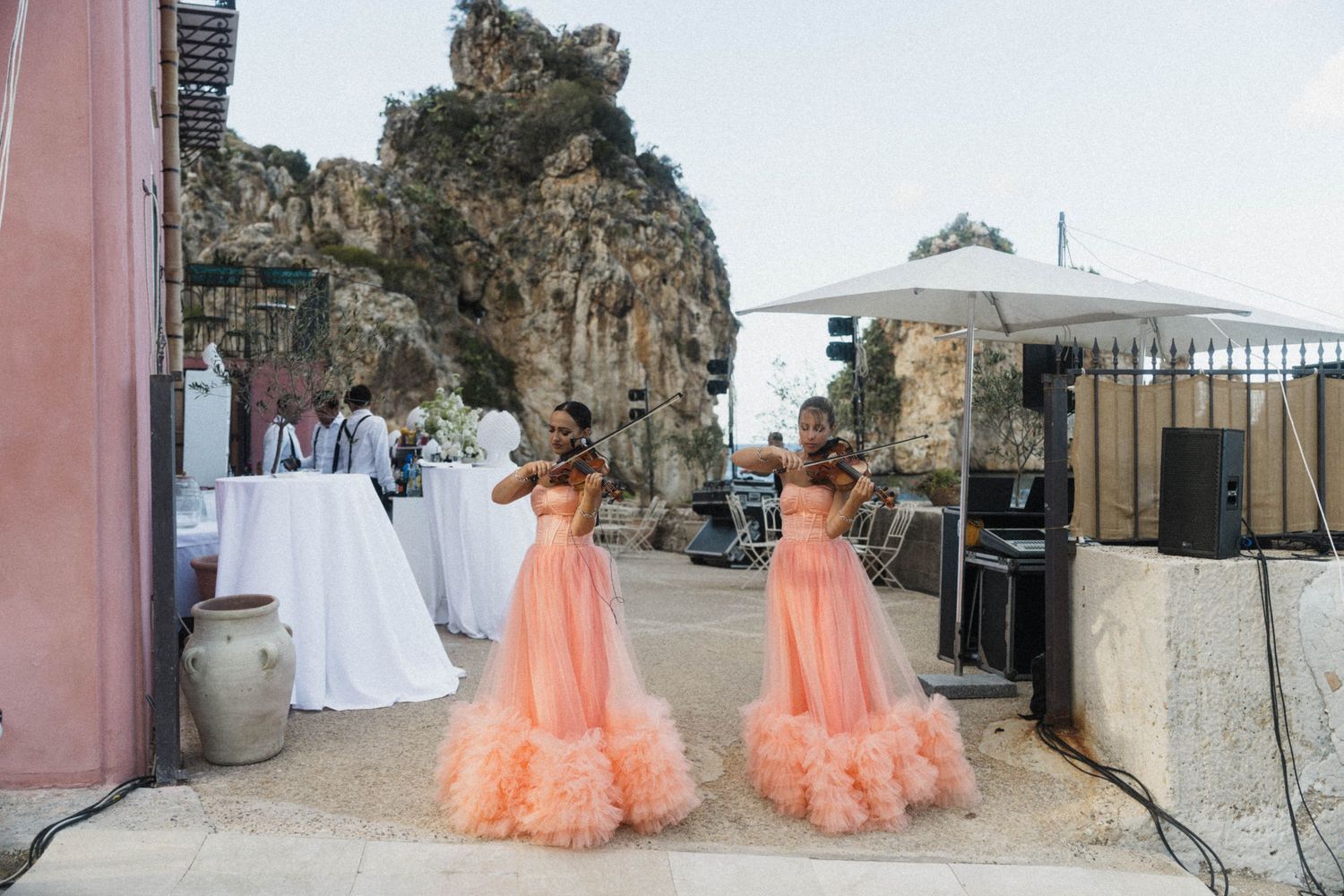 Two violinists in matching peach ruffled gowns performing outdoors at a wedding near dramatic rocky cliffs.