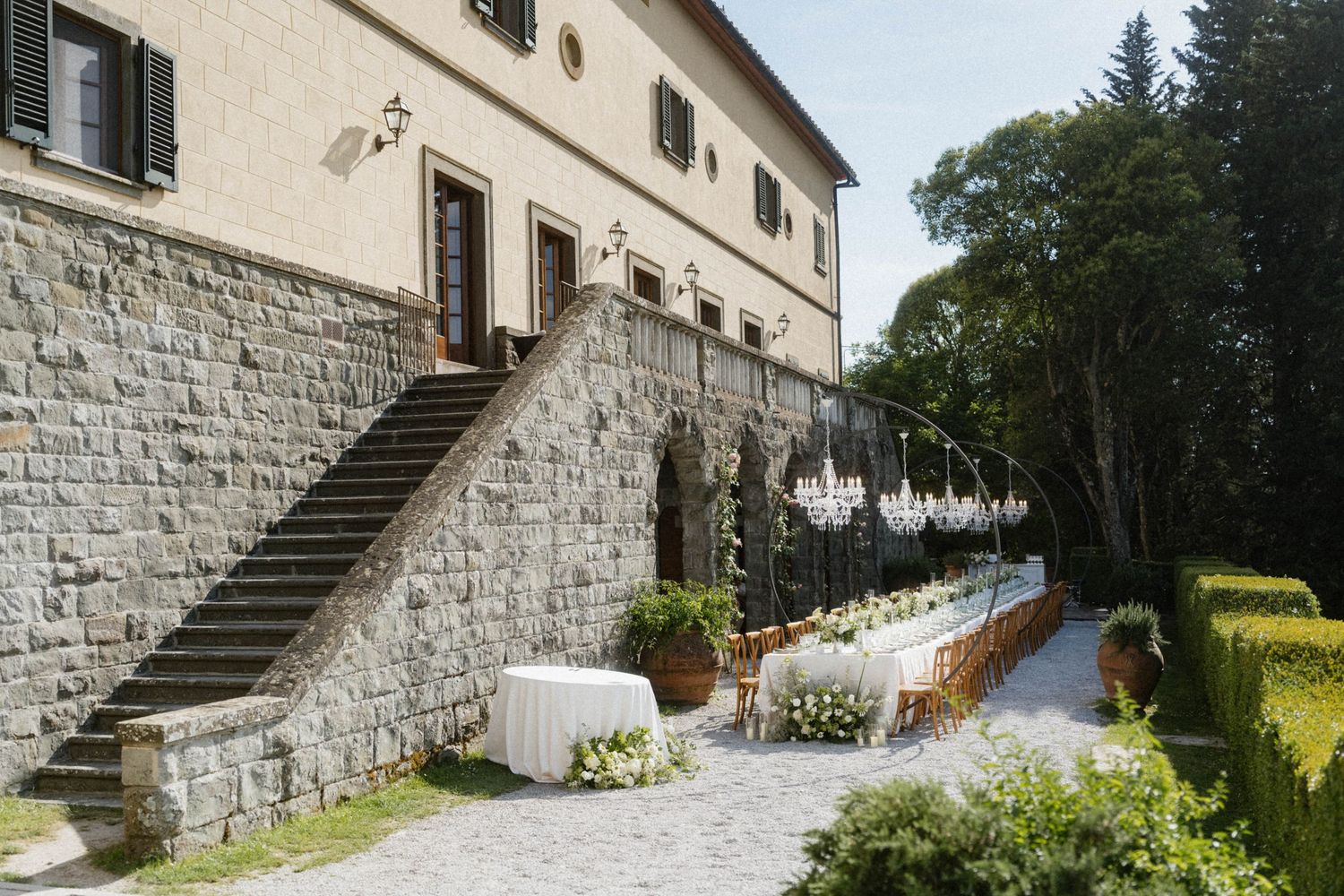 Stunning Tuscan villa wedding venue with stone archways, chandelier arch, long reception table, and manicured hedges.