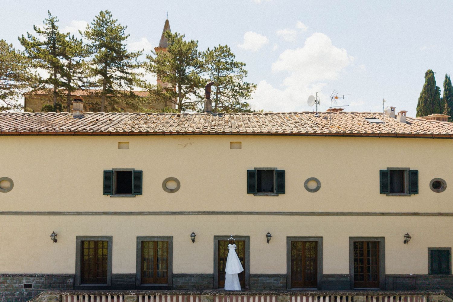 Elegant Italian villa wedding venue with terracotta roof, green shutters, and bride's gown hanging outside.
