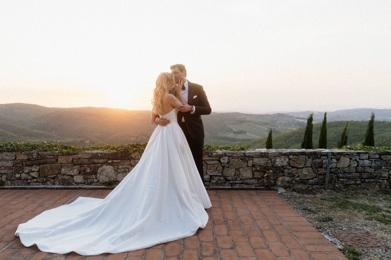 Bride and groom sharing a romantic kiss on a stone terrace with rolling Tuscan hills at sunset.