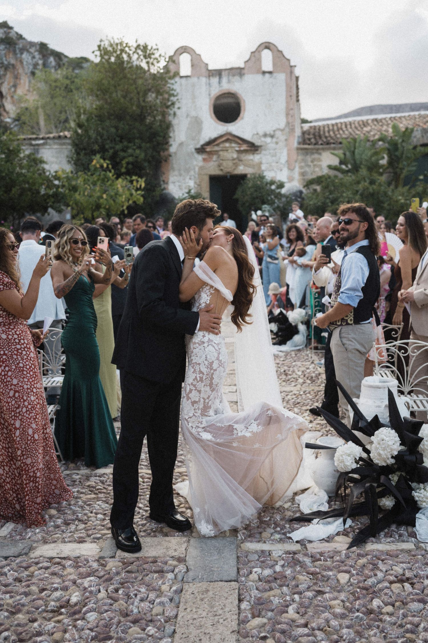 Newlyweds share romantic kiss as wedding guests celebrate on cobblestone path.