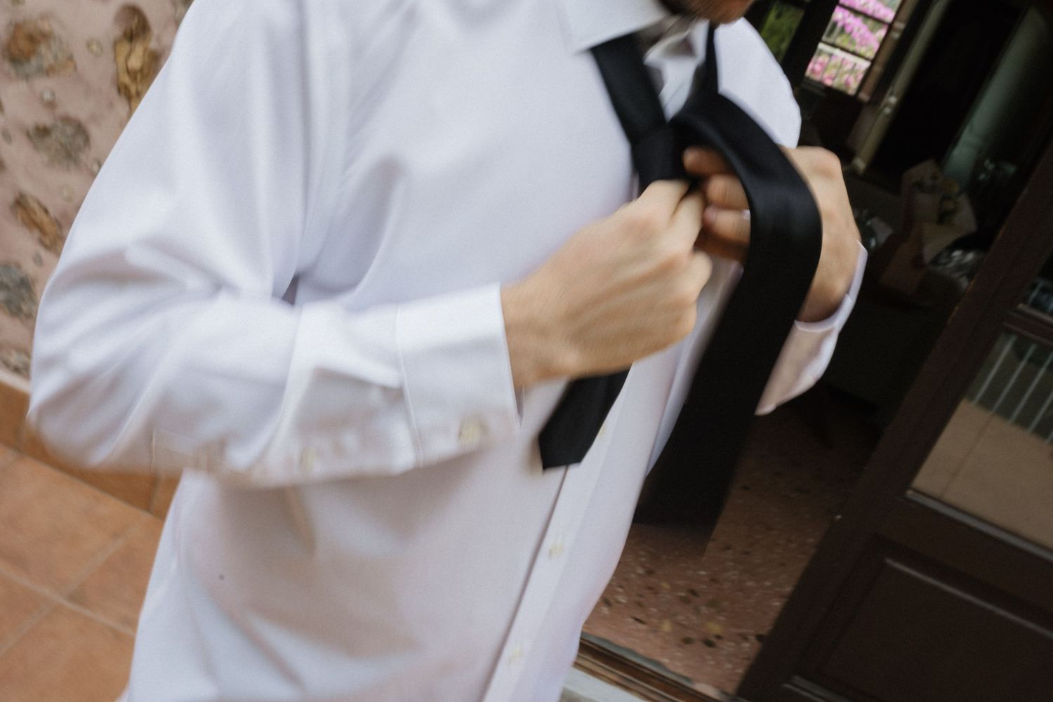 Man in a white dress shirt adjusts a loosely knotted black tie while getting dressed near a doorway.