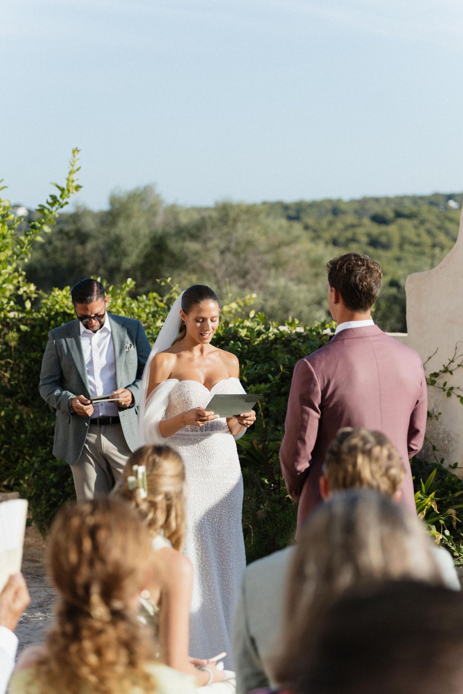 A bride in a white lace gown reads her vows beside the officiant as the groom and flower girls look on outdoors.