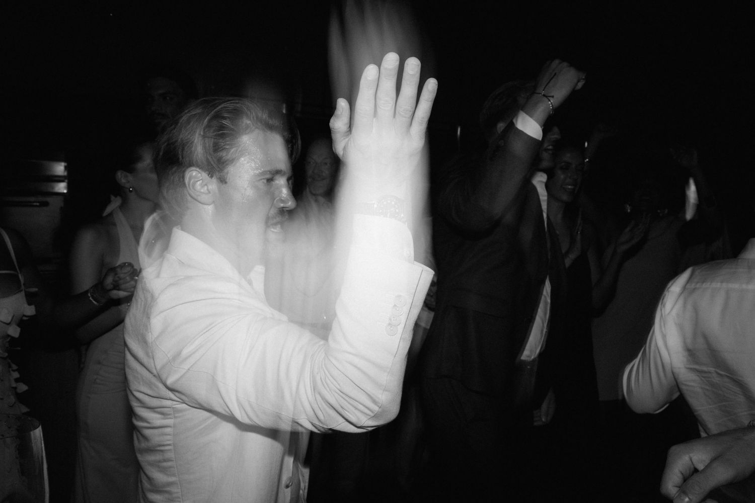Black and white motion-blurred photo of a man in a white shirt raising his hand while dancing at a wedding celebration.