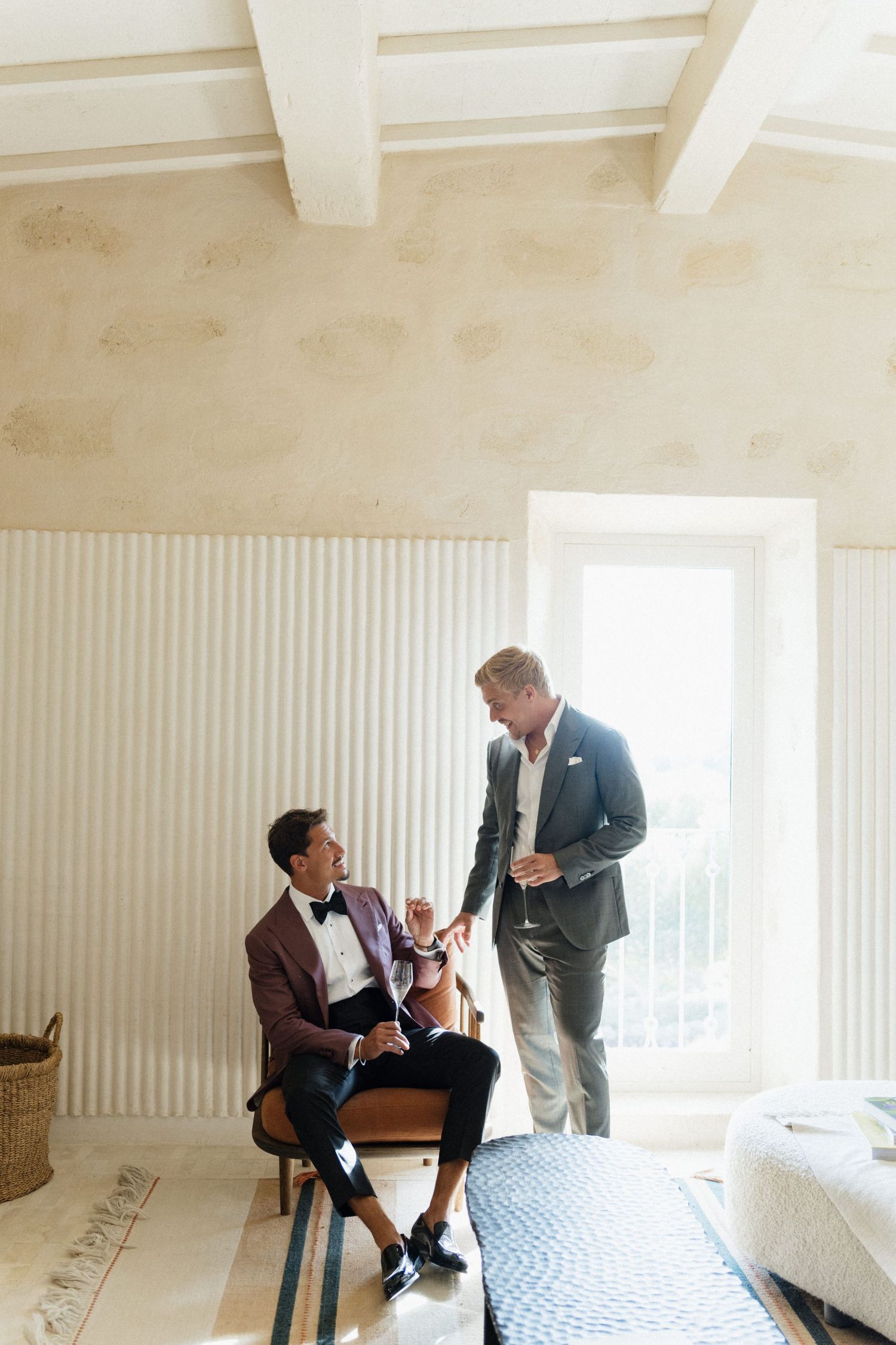 Two men in suits talk near a bright window in a rustic room with white beams, one seated in a burgundy blazer.