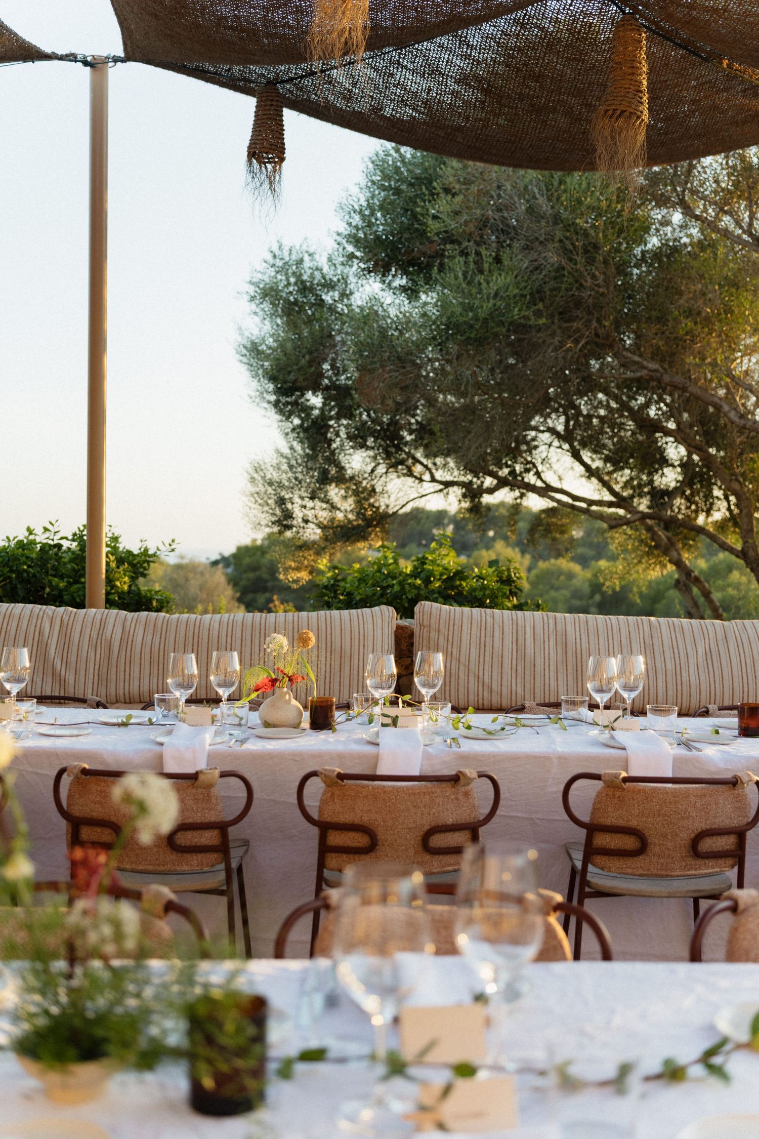 Elegant wedding reception tables with glassware, floral centerpieces and rattan chairs under a woven canopy among olive trees.