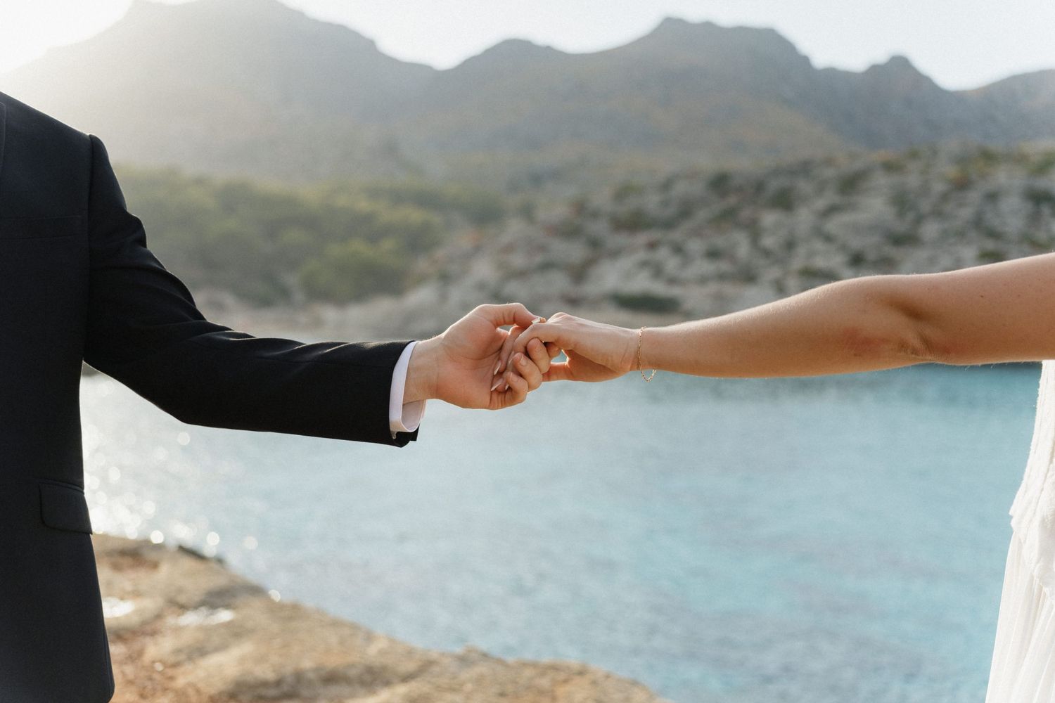 Bride and groom join hands against a blurred turquoise bay and distant mountains on a sunny coastal wedding day.