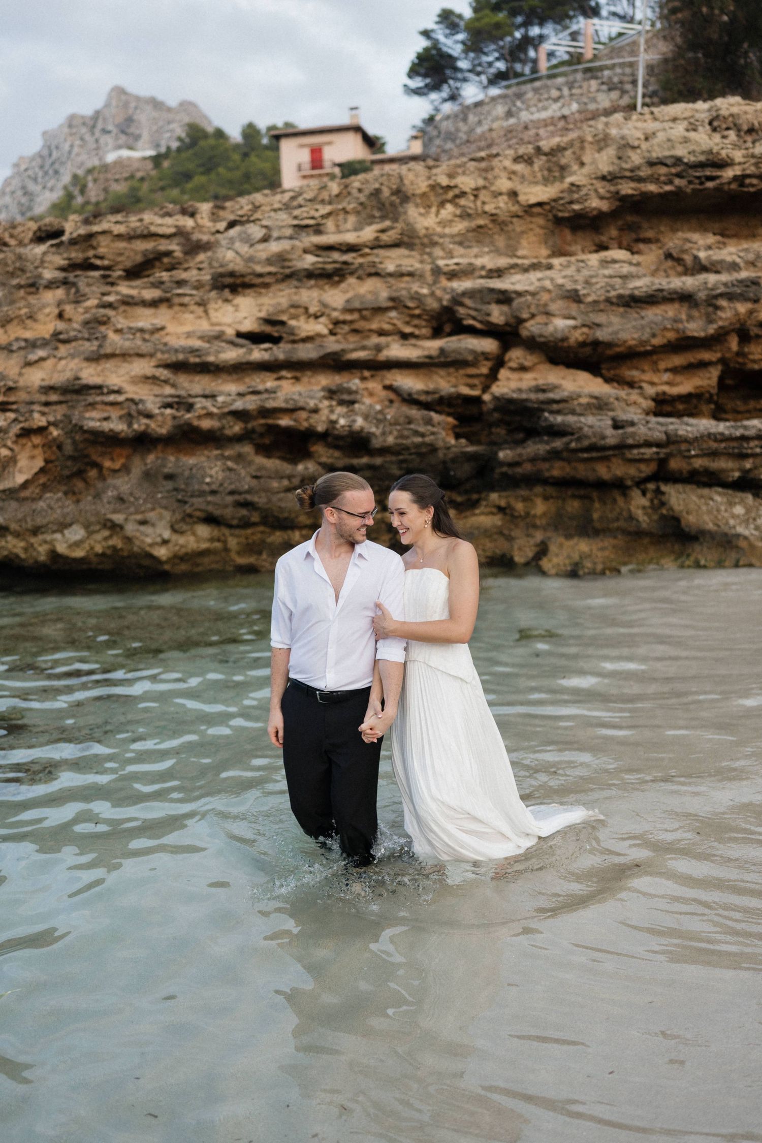 Bride in a white gown and groom in white shirt and black pants walk hand in hand through shallow water beside rocky coastal cliffs.