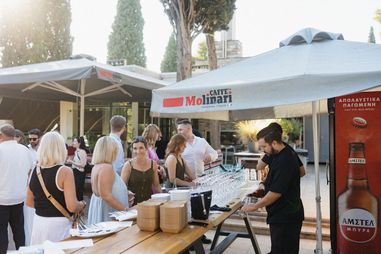 Guests gather at a Caffe Molinari outdoor event with a service bar under a white canopy on a sunny day.