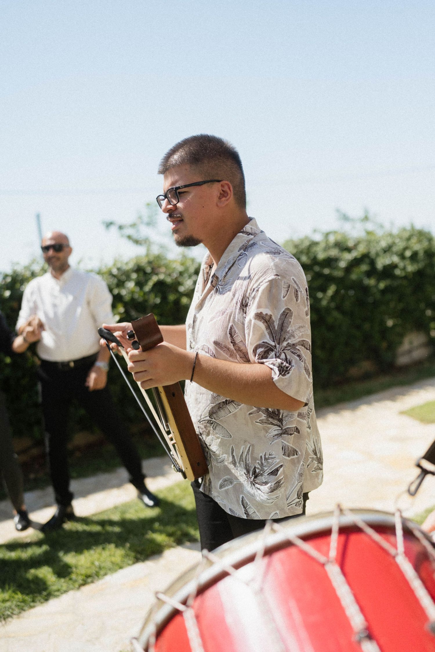 Musician playing guitar outdoors at a wedding celebration with a red drum in the foreground.